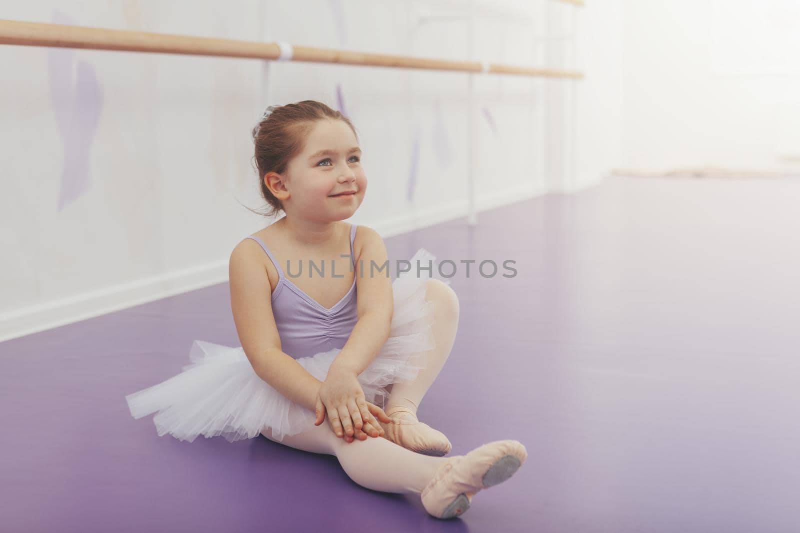 Happy cute little ballerina sitting on the floor at ballet school, smiling looking away dreamily. Adorable little girl dreaming of becoming ballet dancer, exercising at dance studio, copy space