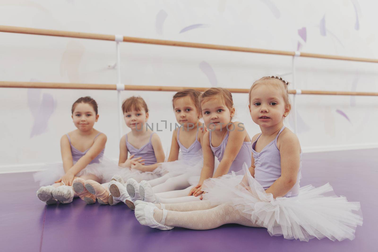 Lovely little girls looking to the camera, sitting on the floor at ballet school. Adorable little ballerinas rest after exercising. Group of little girls dancers wearing leotards relaxing after ballet lesson