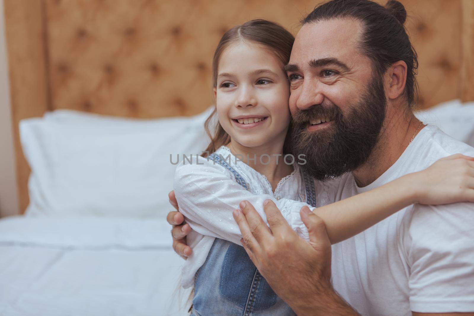 Handsome mature bearded man embracing his little daughter, smiling looking away joyfully. Happy little girl hugging her dad at home, copy space