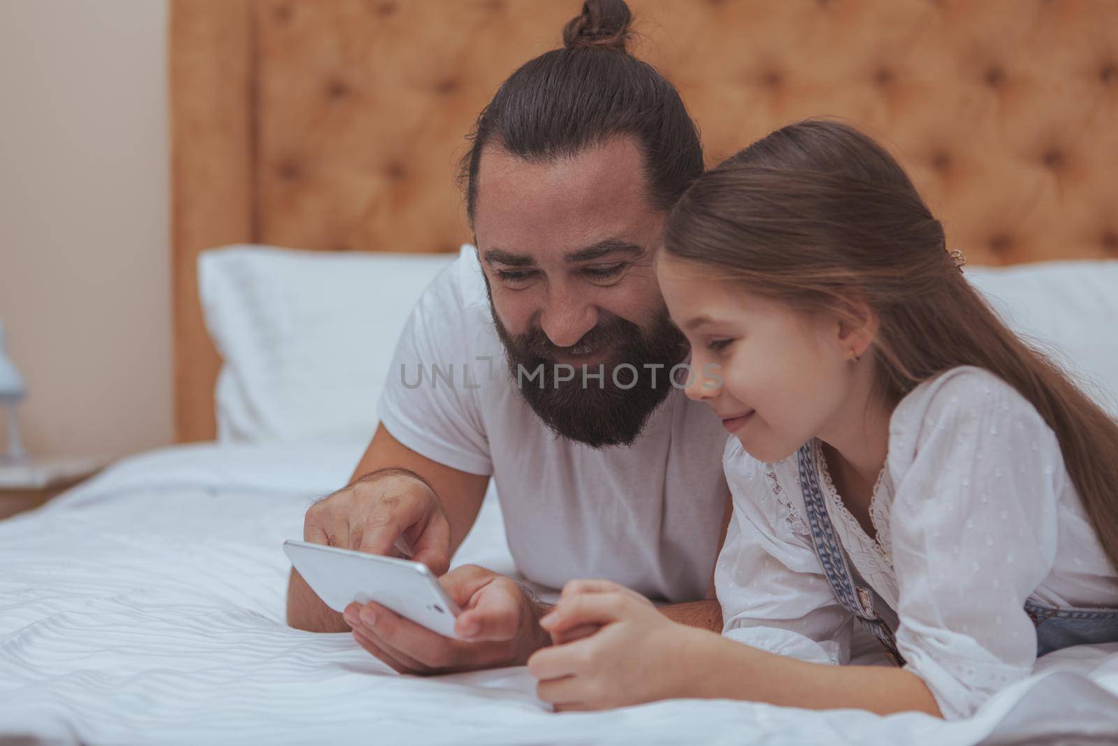 Cute happy little girl and her father using smart phone together, relaxing at home. Handsome bearded man showing his little daughter how to use mobile phone, copy space