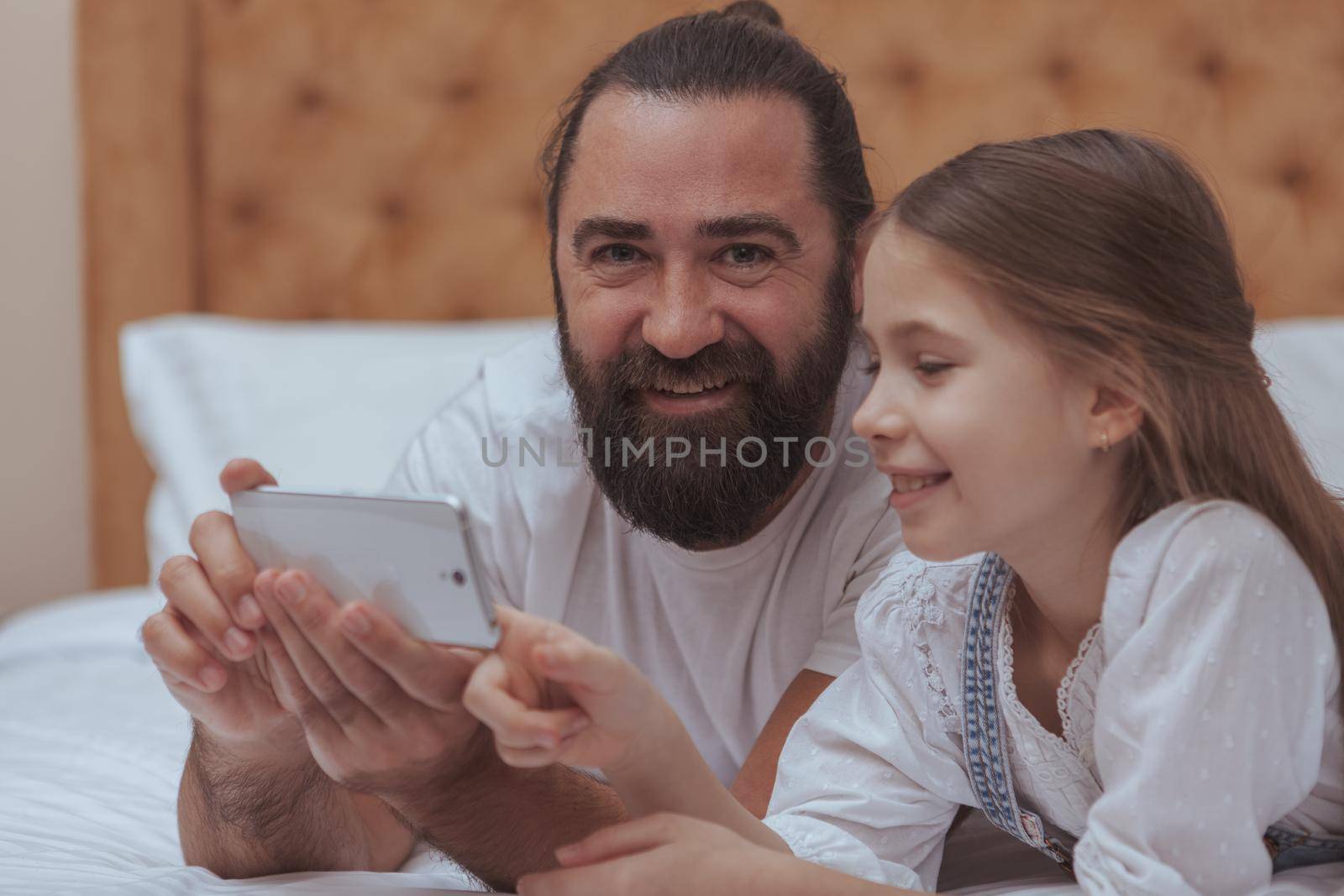 Handsome bearded man smiling to the camera, while using smart phone with his adorable daughter. Cute little girl smiling, using mobile phone with her dad. Online, internet, gadget concept