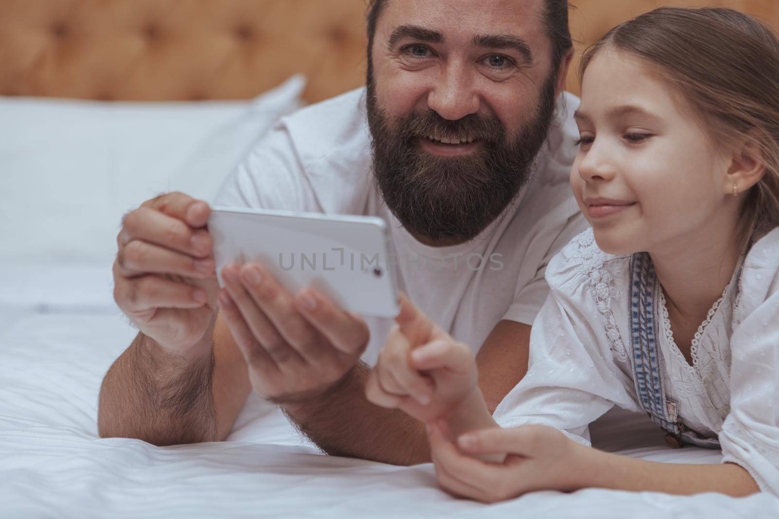 Cropped close up of a handsome mature bearded man smiling to the camera, using smart phone with his little daughter. Adorable little girl learning using mobile phone with her father