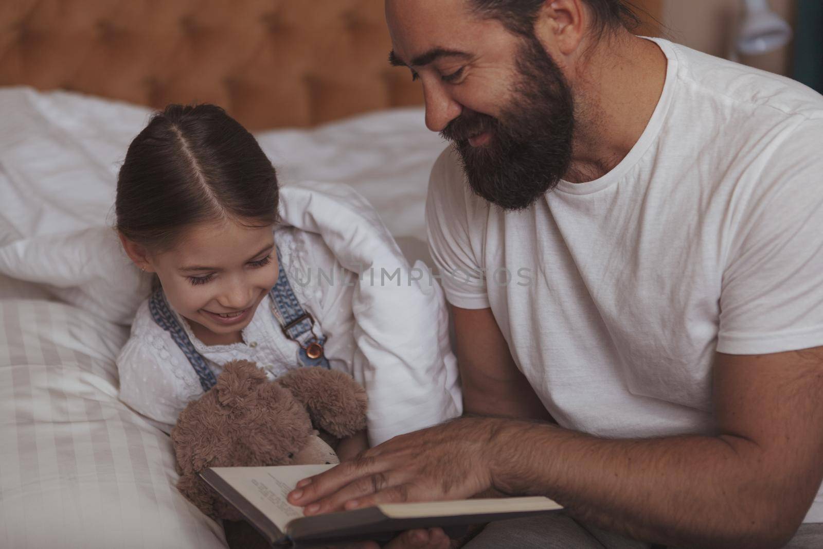 Happy father and daughter enjoy reading a book together at night before sleep. Bearded cheerful man reading fairytales to his adorable daughter, resting together at home in the evening