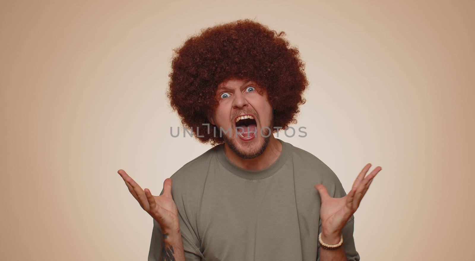 Man with lush brown hairstyle screams from stress tension problems feels horror hopelessness fear surprise shock expresses gestures rage. Young guy boy isolated alone on beige studio wall background