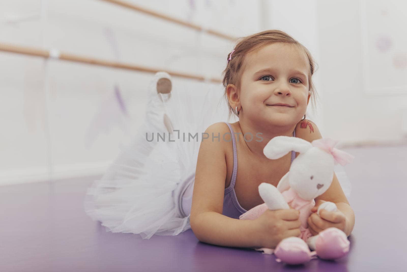 Beautiful little happy girl wearing leotard and tutu lying on the floor at dance school. Cute cheerful little ballerina at ballet studio, copy space. Ambition, dreaming, achievement concept