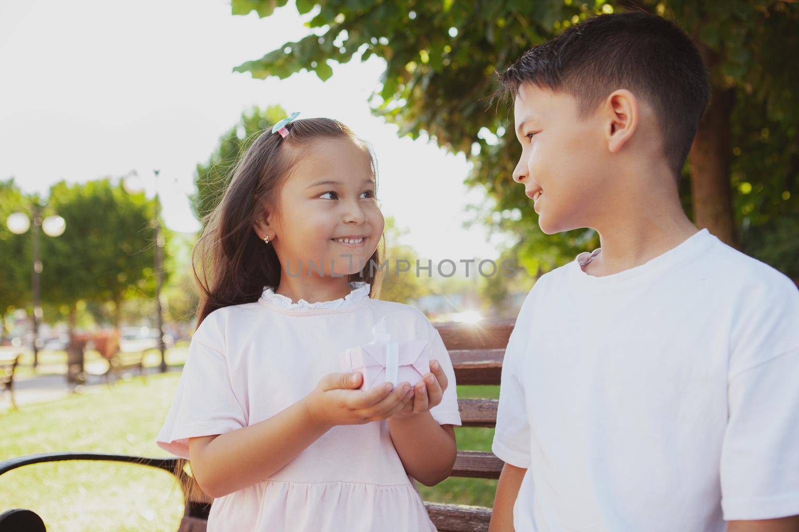 Little adorable Asian girl giving small present to her brother outdoors. Cheerful little girl holding gift box, congratulating her brother. Kids exchange presents in the park