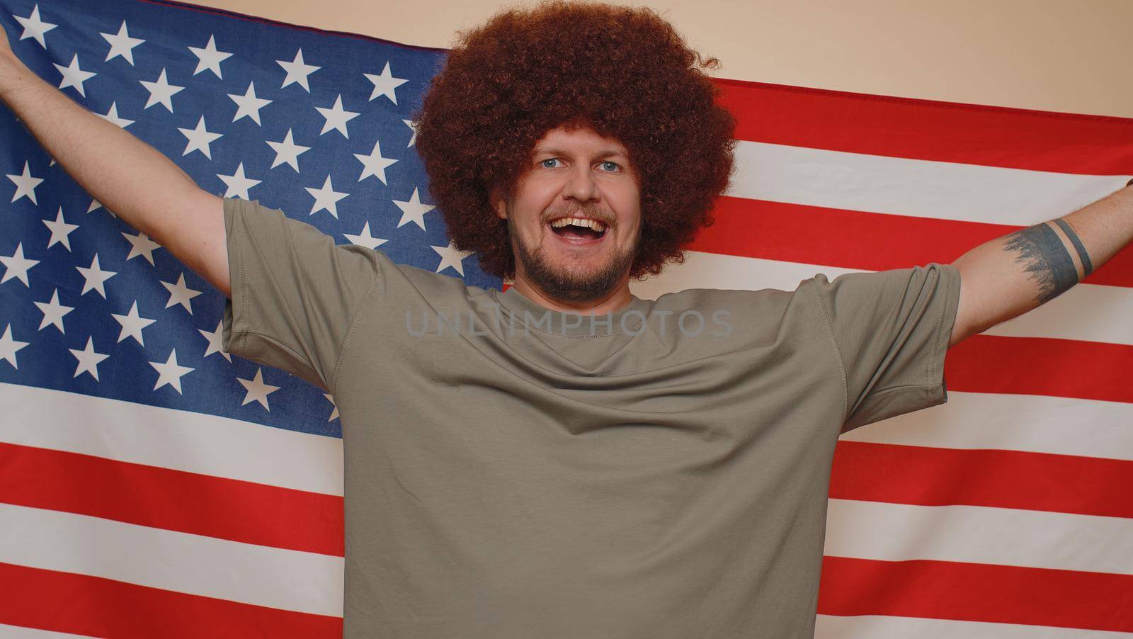 Handsome bearded man with lush hairstyle waving and wrapping in American USA flag, celebrating, human rights and freedoms. Independence day. Young adult guy boy isolated on beige studio background
