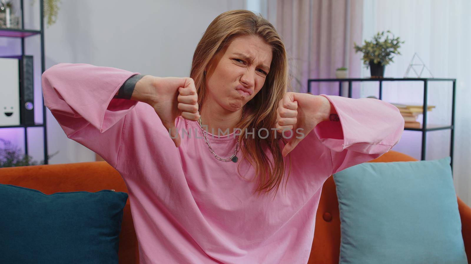 Dislike. Upset girl showing thumbs down sign gesture, expressing discontent, disapproval, dissatisfied bad work at modern home apartment indoors. Displeased young woman in living room sitting on sofa