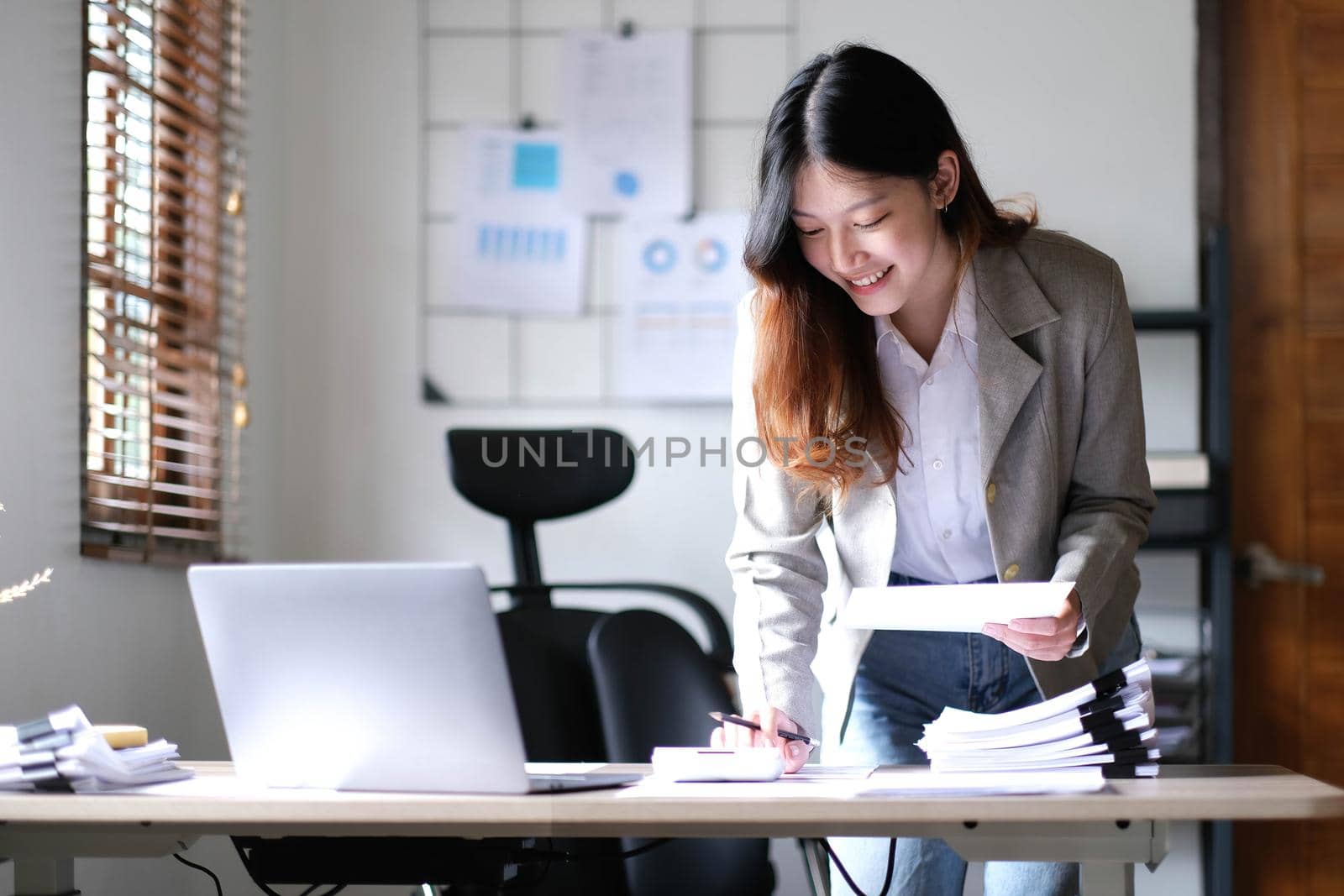 Beautiful Asian businesswoman standing and working on laptop with smiling face while working in office..