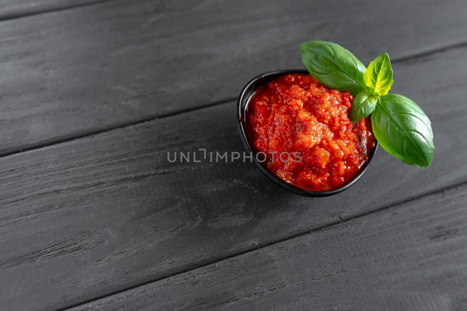 Ajvar in a black pial with basil on a wooden black background. Sweet bell pepper paste.