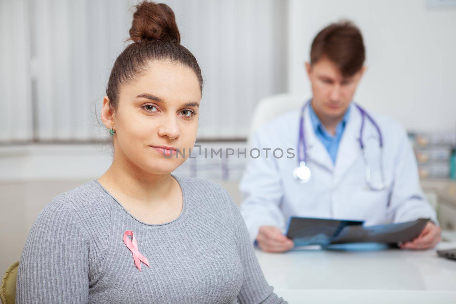 Charming yong woman with breast cancer pink ribbon on her chest looking to the camera confidently, doctor examining x-ray scans on background