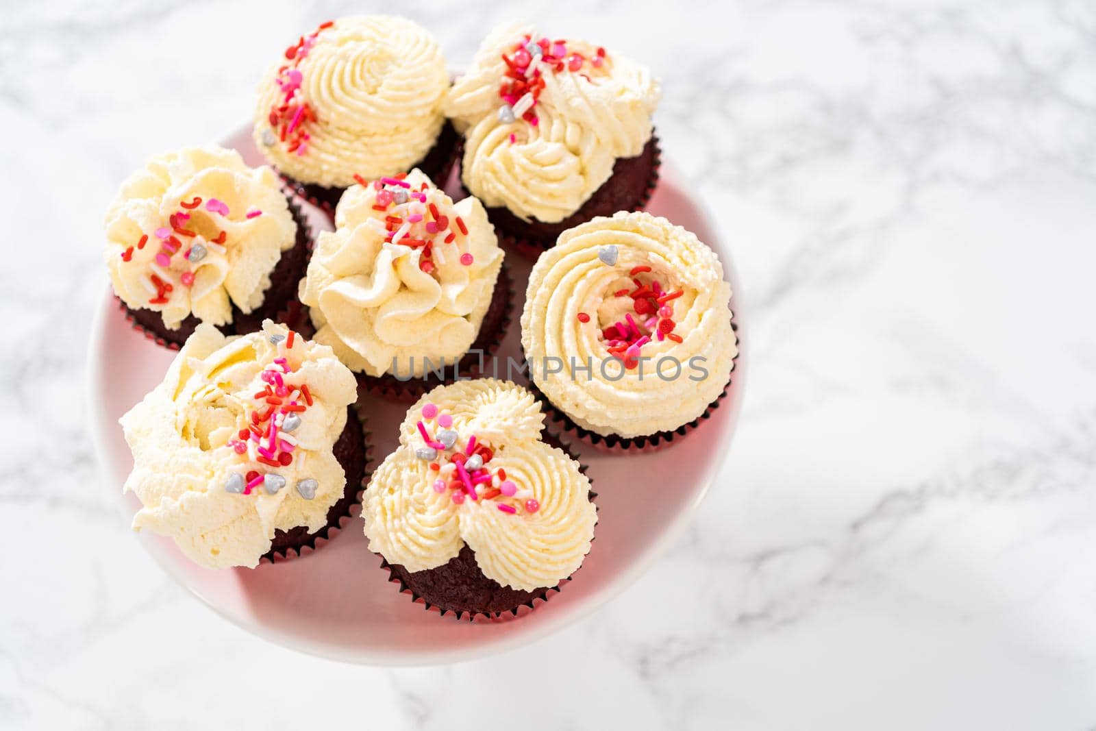 Freshly baked velvet cupcakes with white chocolate ganache frosting decorated with sprinkles.