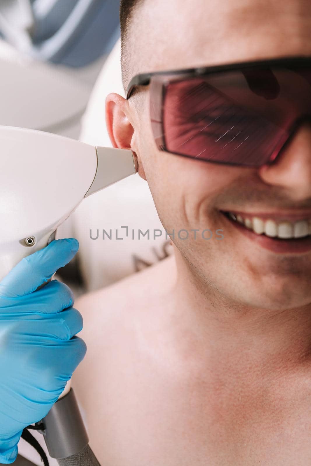 Cropped close up of a man smiling, getting laser hair removal treatment on his ear. Beautician removing hair on the ear of a male client, using laser