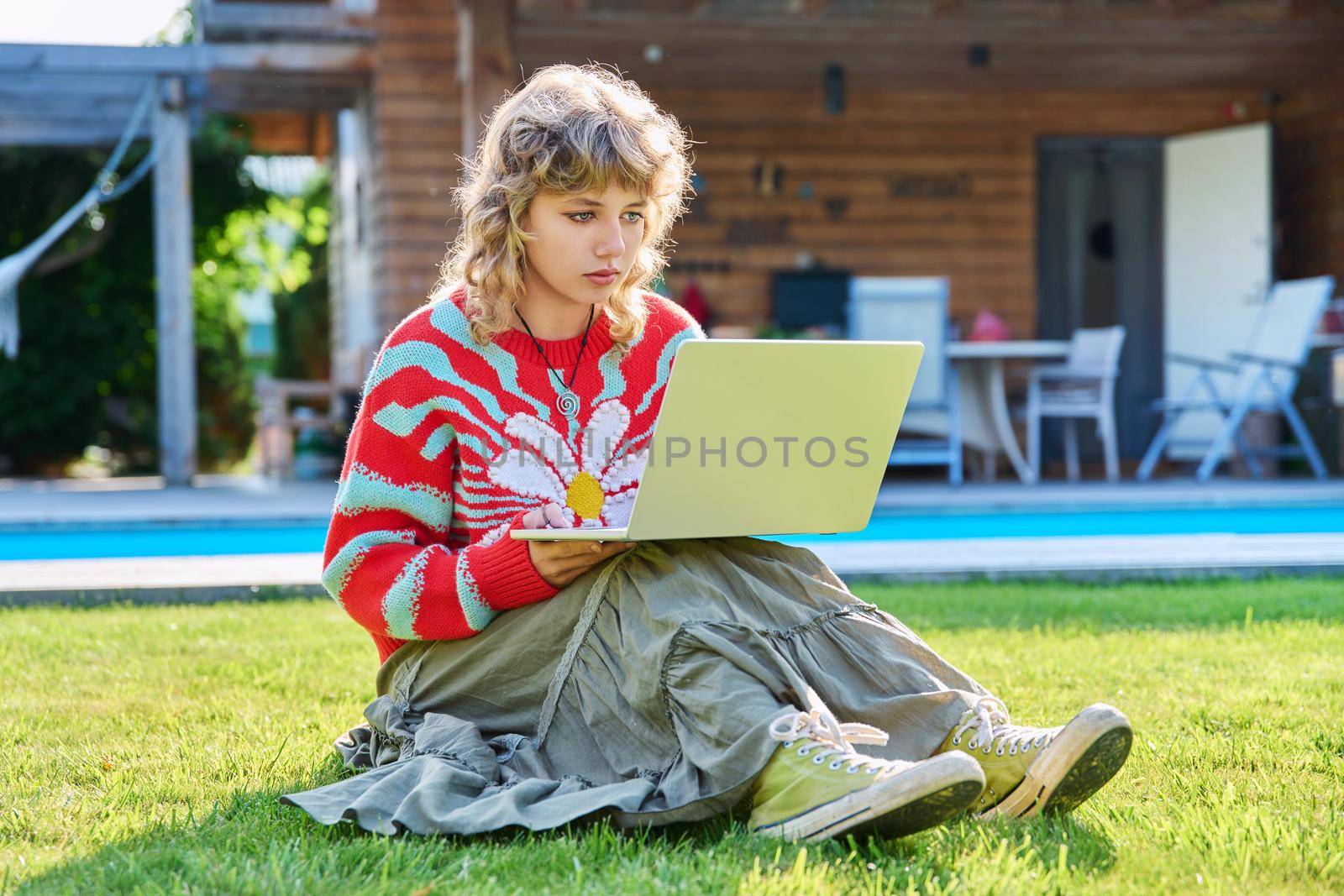 Teenage girl 17 years old sitting on lawn in backyard, using laptop for studying leisure. Adolescence, students, high school, technology, lifestyle, youth concept