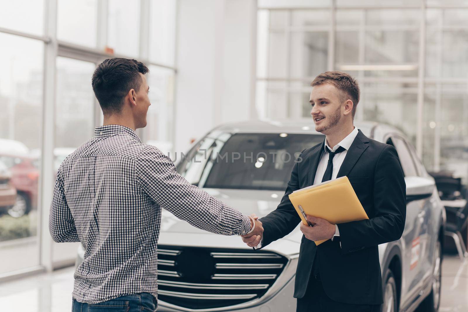 Professional car salesman smiling, shaking hands with his male customer in front of a new automobile. Man buying new car at the salon, shaking hands with salesperson. Service, occupation concept