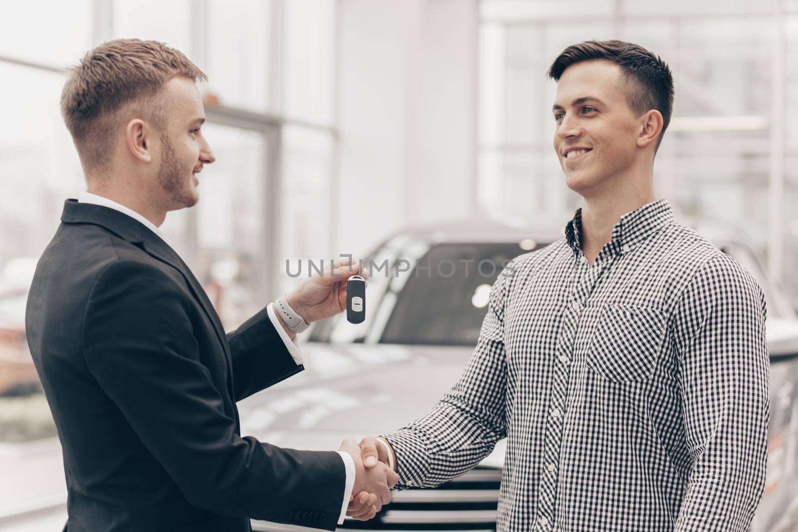 Happy handsome man receiving car keys to his new automobile from car salesman at the dealership. Professional auto seller shaking hands with his customer, handing him keys to a new vehicle. Buying car