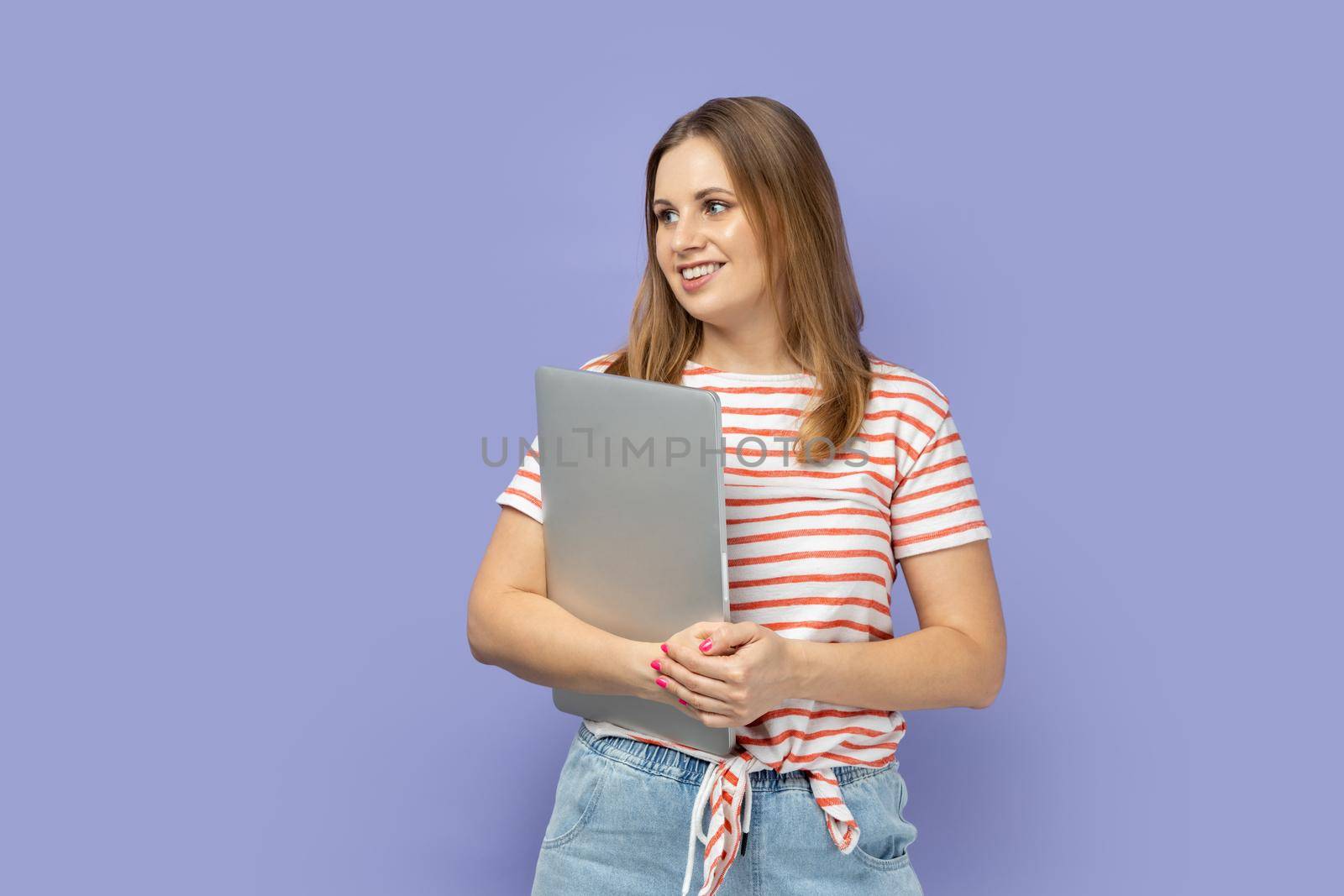 Portrait of beautiful positive blond woman wearing striped T-shirt standing with closed laptop after finishing work, looking away. Indoor studio shot isolated on purple background.