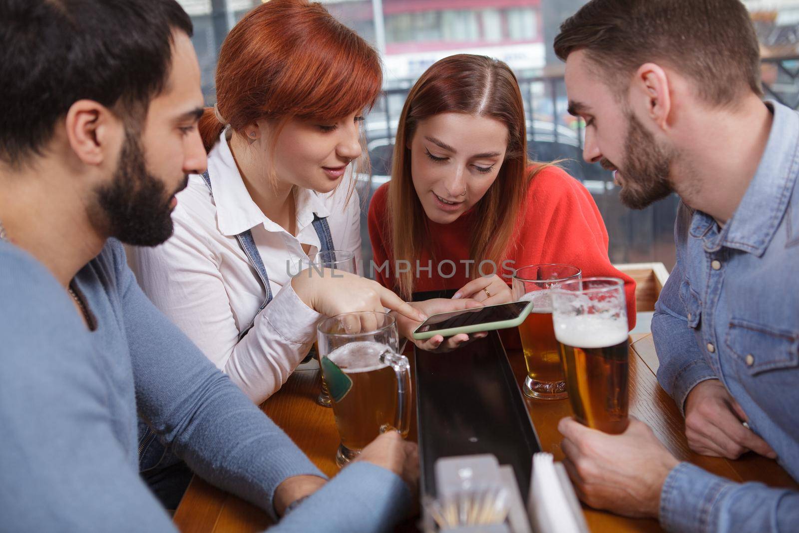 Group of friends using smart phone while drinking beer together. Young beautiful woman showing something online to her friends, using her phone