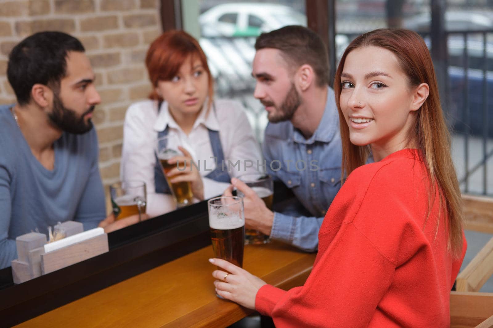 Attractive young woman smiling to the camera while drinking beer at the pub with her friends. Festivity, celebration concept