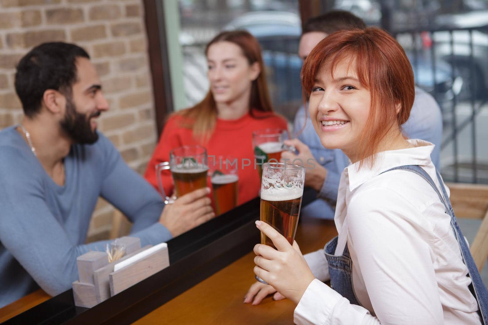 Happy beautiful woman smiling to the camera, holding glass of delicious craft beer, her friends talking on background