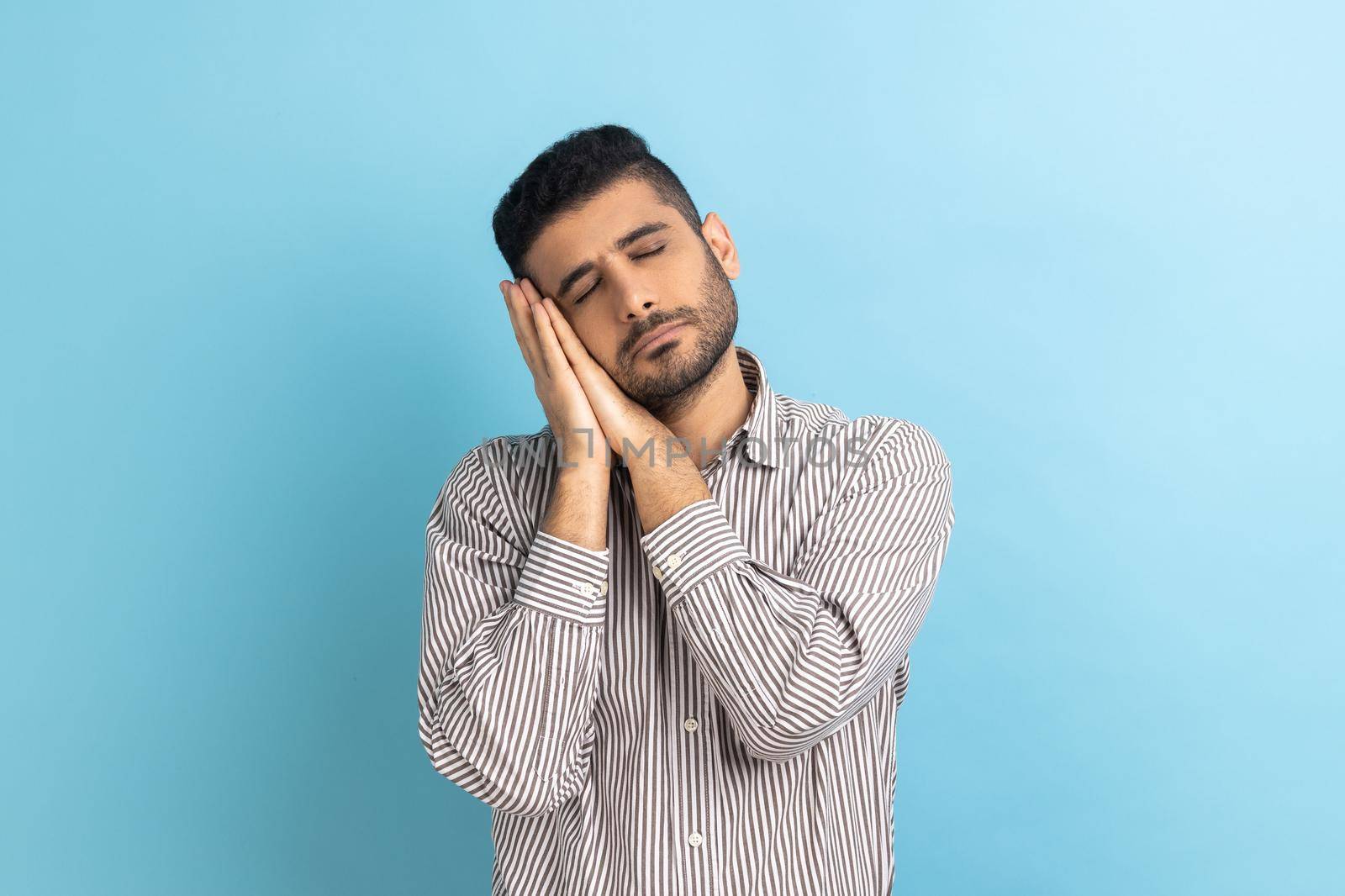 Portrait of handsome bearded man sleeping laying down on her palms, having comfortable nap and resting, dozing off, wearing striped shirt. Indoor studio shot isolated on blue background.