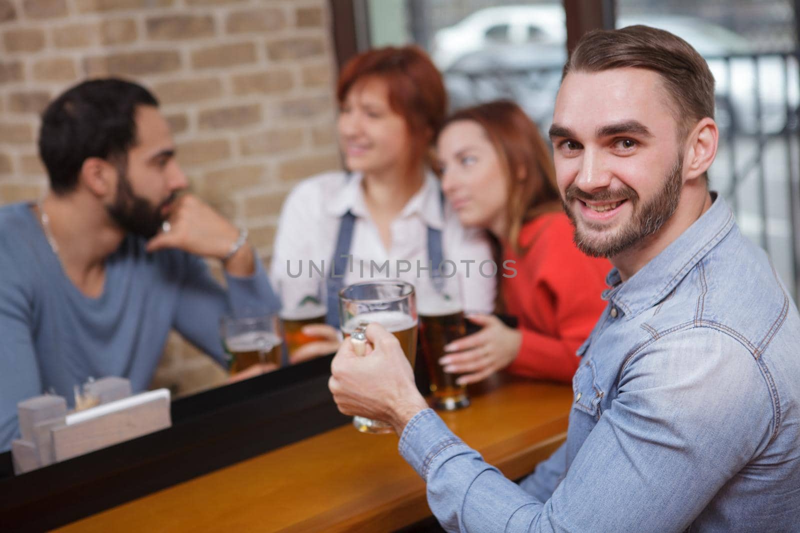 Handsome bearded man smiling to the camera, drinking beer with his friends at the pub