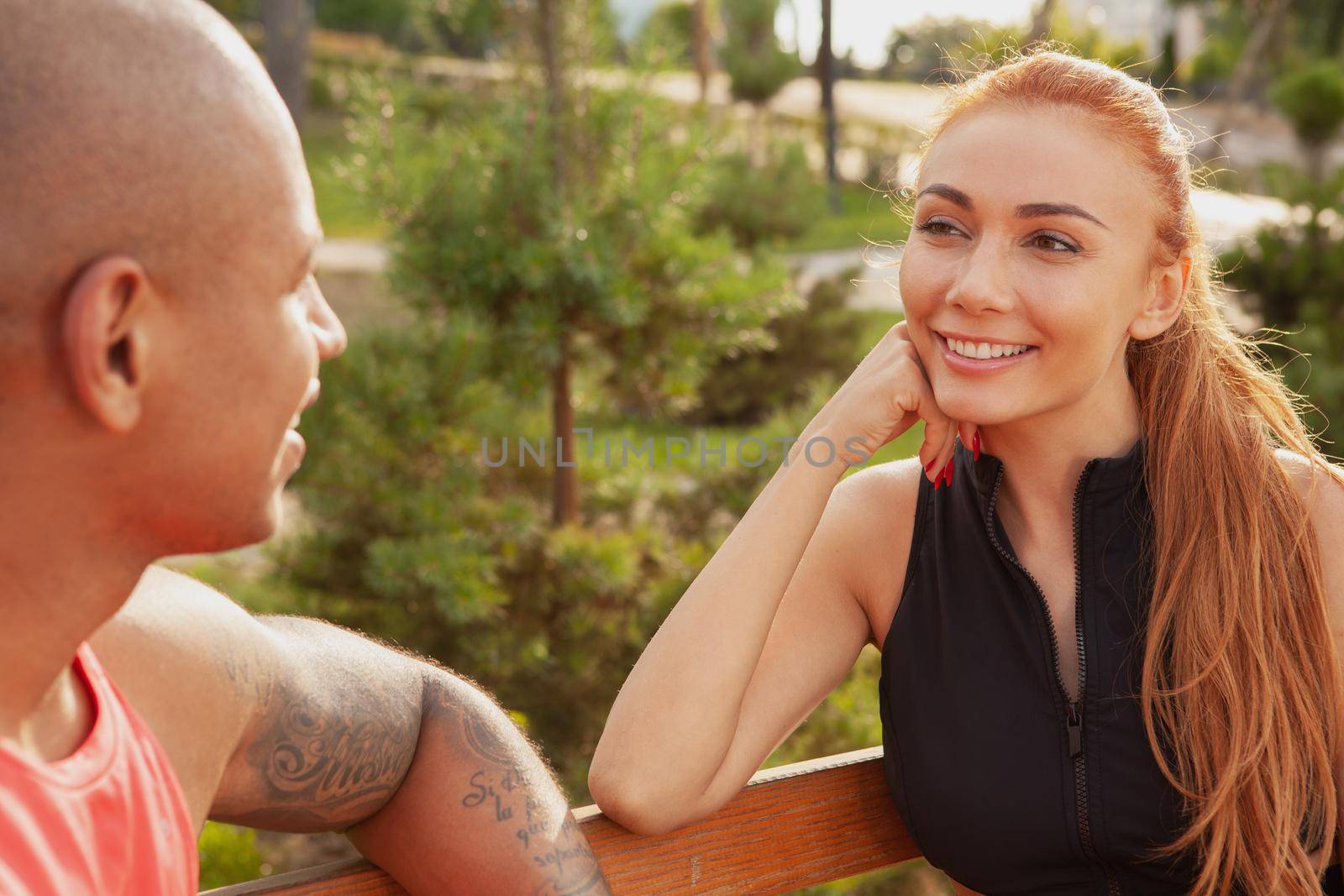 Beautiful happy woman smiling joyfully at her boyfriend, resting together outdoors after working out. Charming couple talking, sitting on the bench