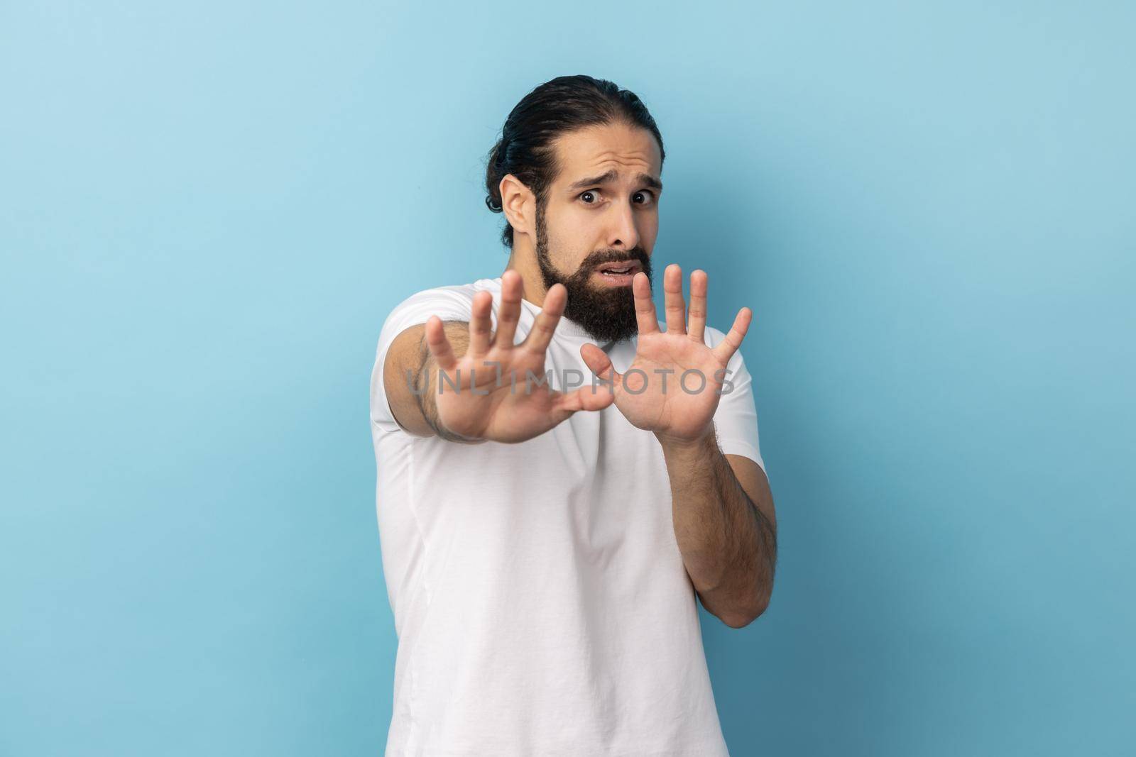 Portrait of nervous embarrassed man with beard wearing white T-shirt feels scared reacts on something looks anxious, having anxiety or being troubled. Indoor studio shot isolated on blue background.
