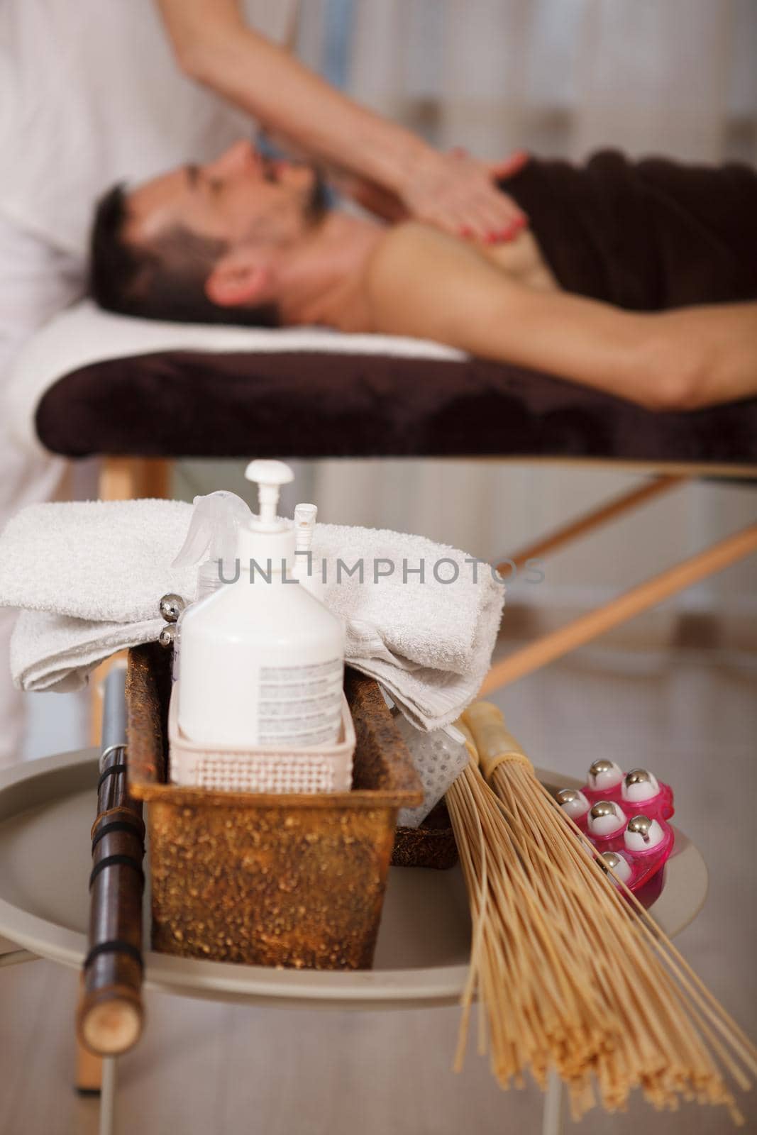 Vertical close up of skin moisturizer bottle and massage tools, man getting professional spa massage on background