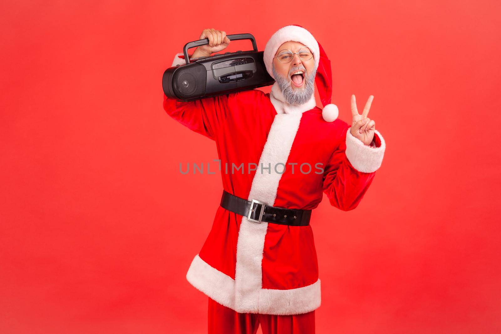 Positive funny elderly man with gray beard wearing santa claus costume standing and holding tape recorder showing v sign, celebrating Christmas party. Indoor studio shot isolated on red background.