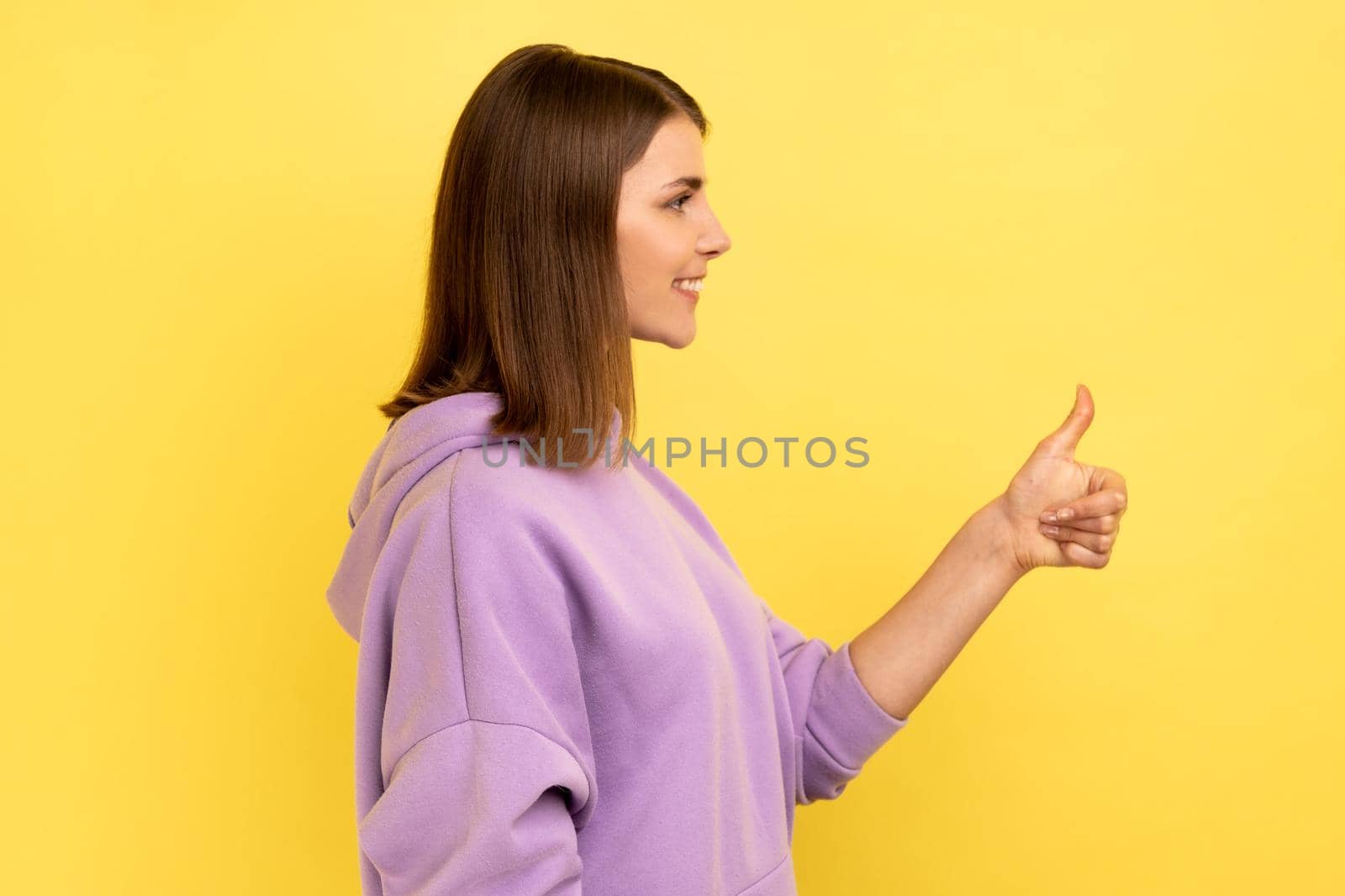Side view of woman looking ahead with toothy smile, showing thumb up, approval sign, satisfied with service, good feedback, wearing purple hoodie. Indoor studio shot isolated on yellow background.