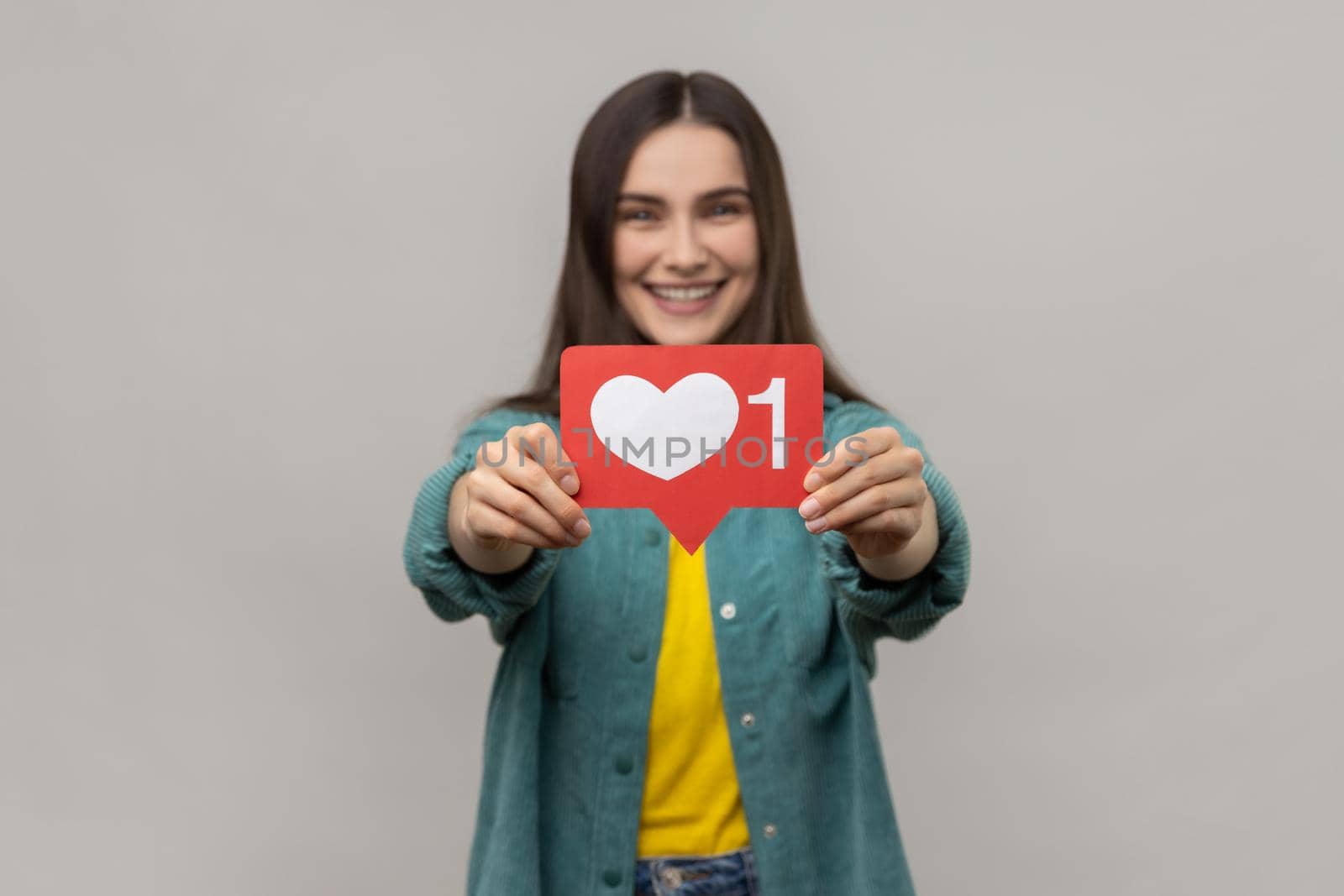 Satisfied woman holding out heart icon, recommending to follow and share blog with interesting content, wearing casual style jacket. Indoor studio shot isolated on gray background.