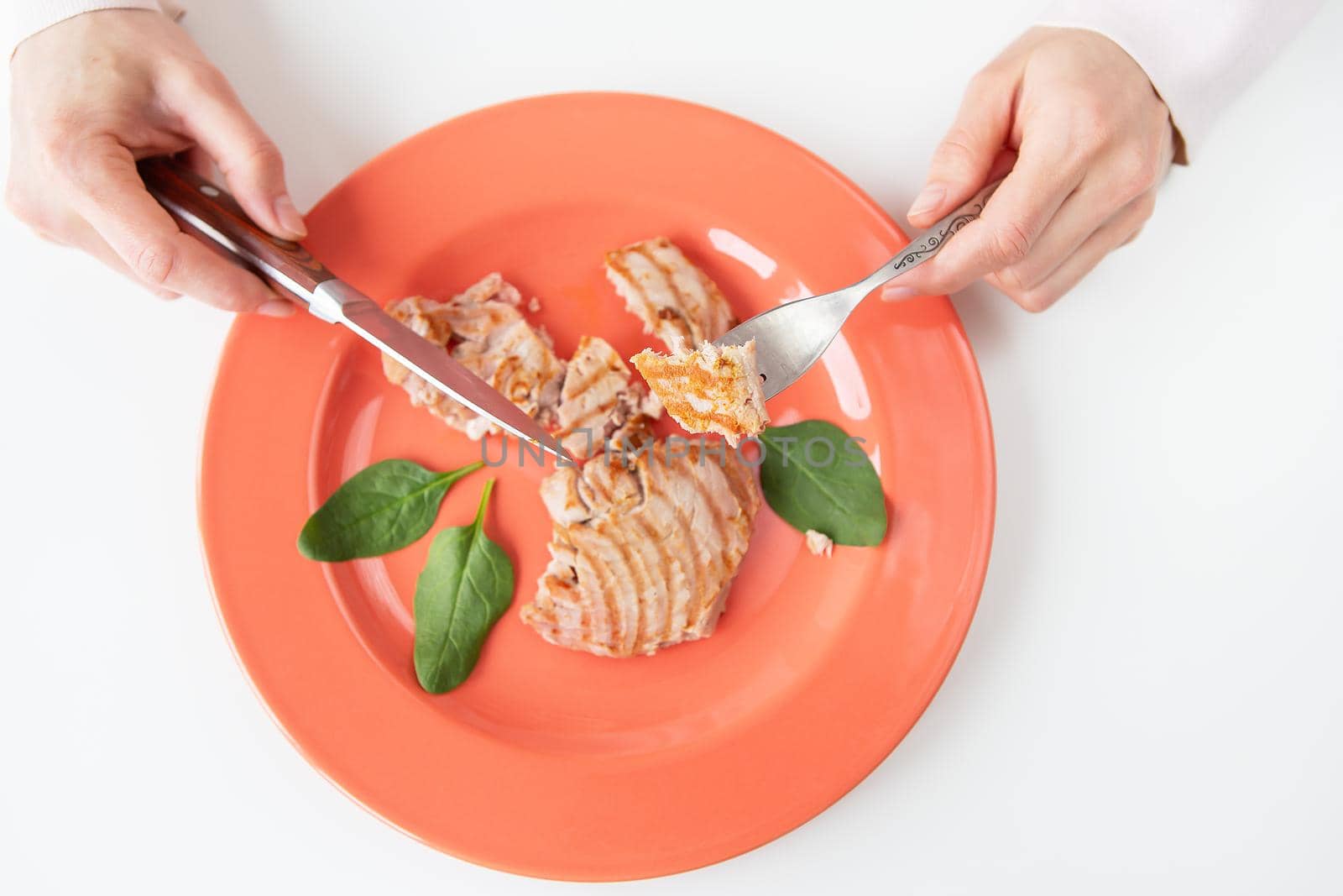 Close-up shot of a juicy delicious grilled tuna steak on a bright coral plate. Delicious and healthy food, proper nutrition. The girl is holding a fork and knife. View from above. by sfinks