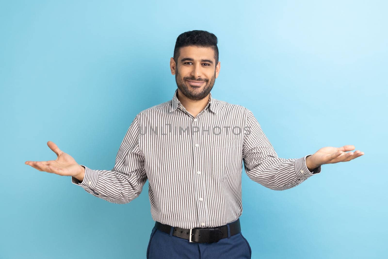 Open hearted generous businessman with beard sharing opening hands looking at camera with kind smile, greeting and regaling, wearing striped shirt. Indoor studio shot isolated on blue background.