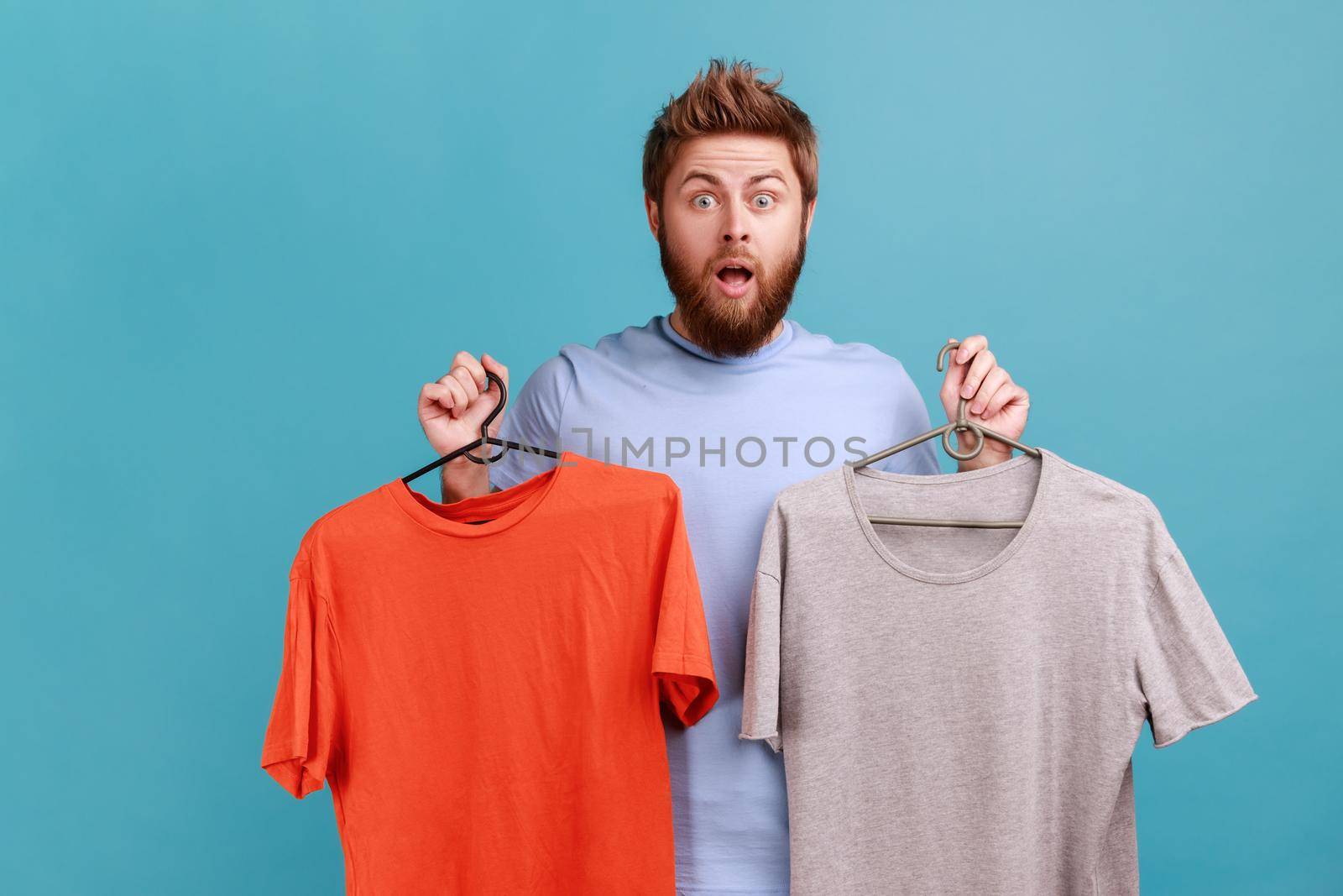 Portrait of surprised astonished handsome bearded man holding two hangers with gray and orange T-shirts, expressing shock, keeps mouth opened. Indoor studio shot isolated on blue background.