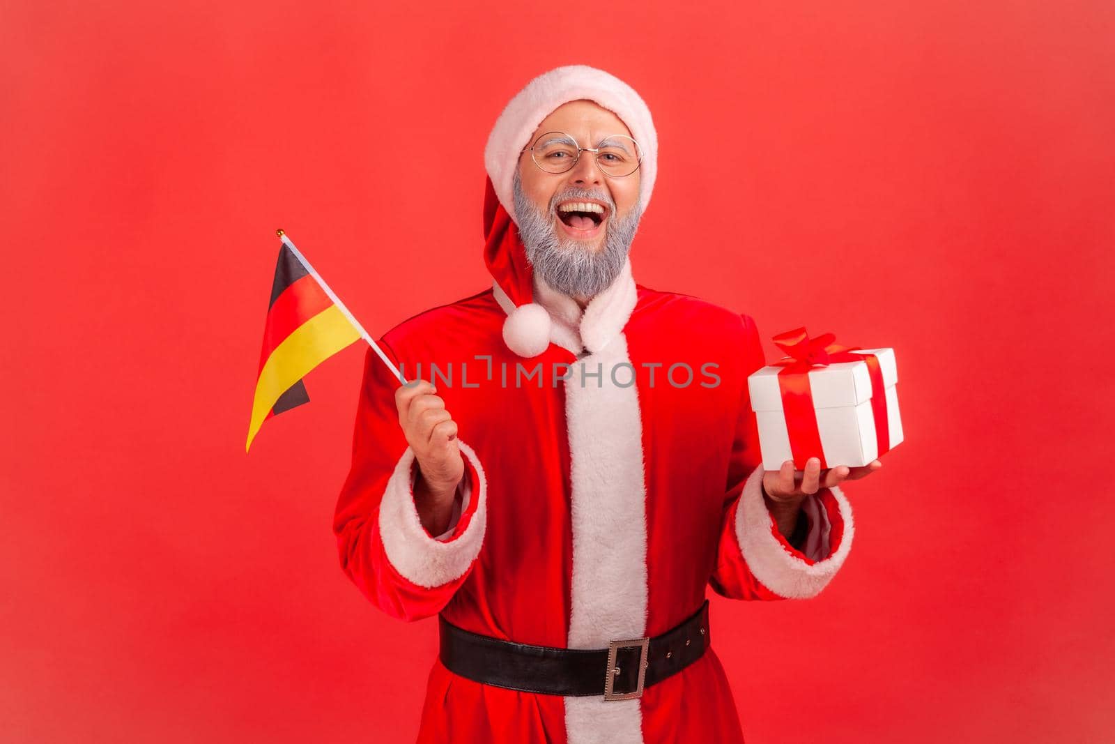 Portrait of happy excited elderly man with gray beard wearing santa claus costume holding German flag and box with Christmas present. Indoor studio shot isolated on red background.