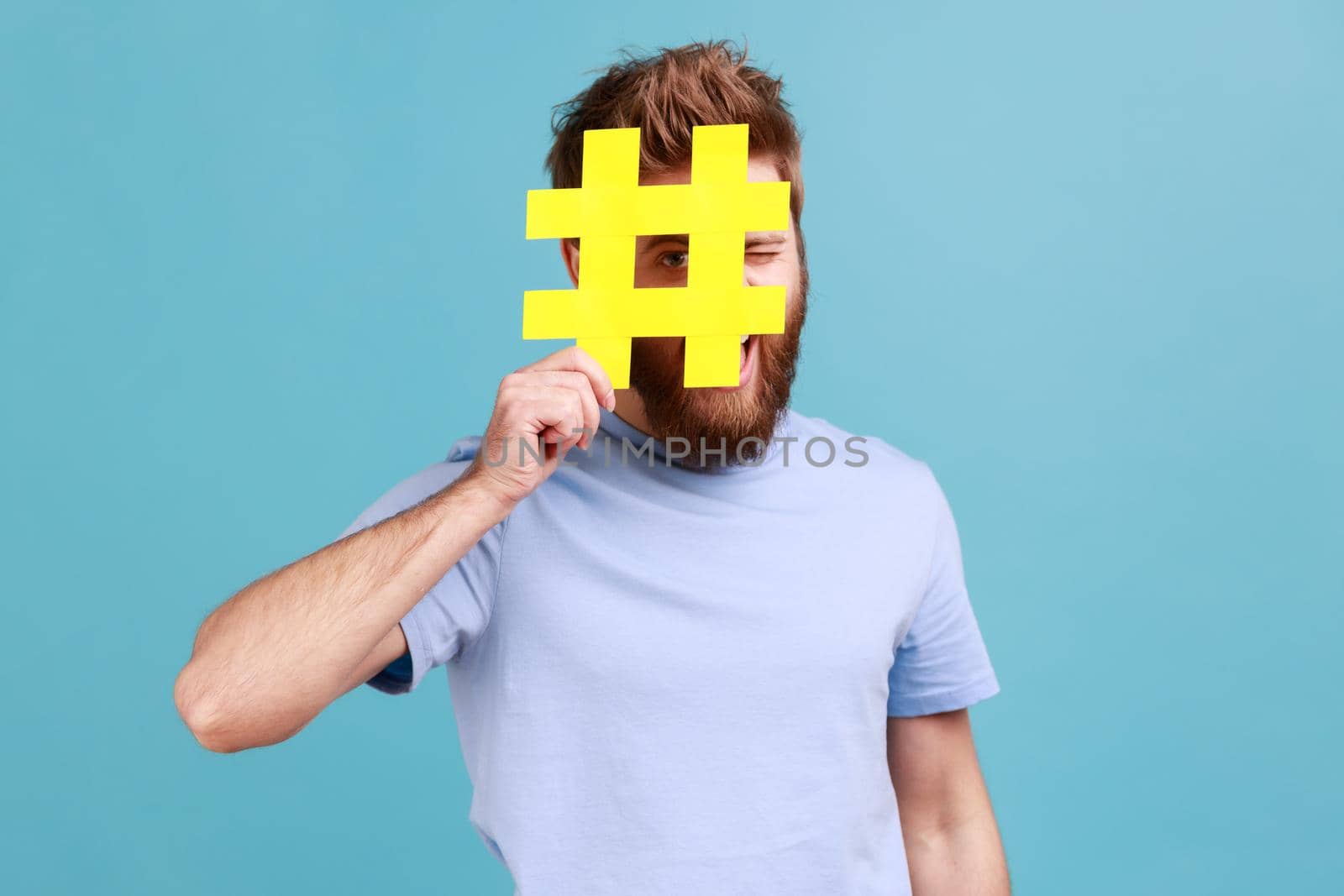 Portrait of bearded man hiding face behind yellow hashtag sign, calls on to tag topics on websites, giving recommendations, peeping. Indoor studio shot isolated on blue background.