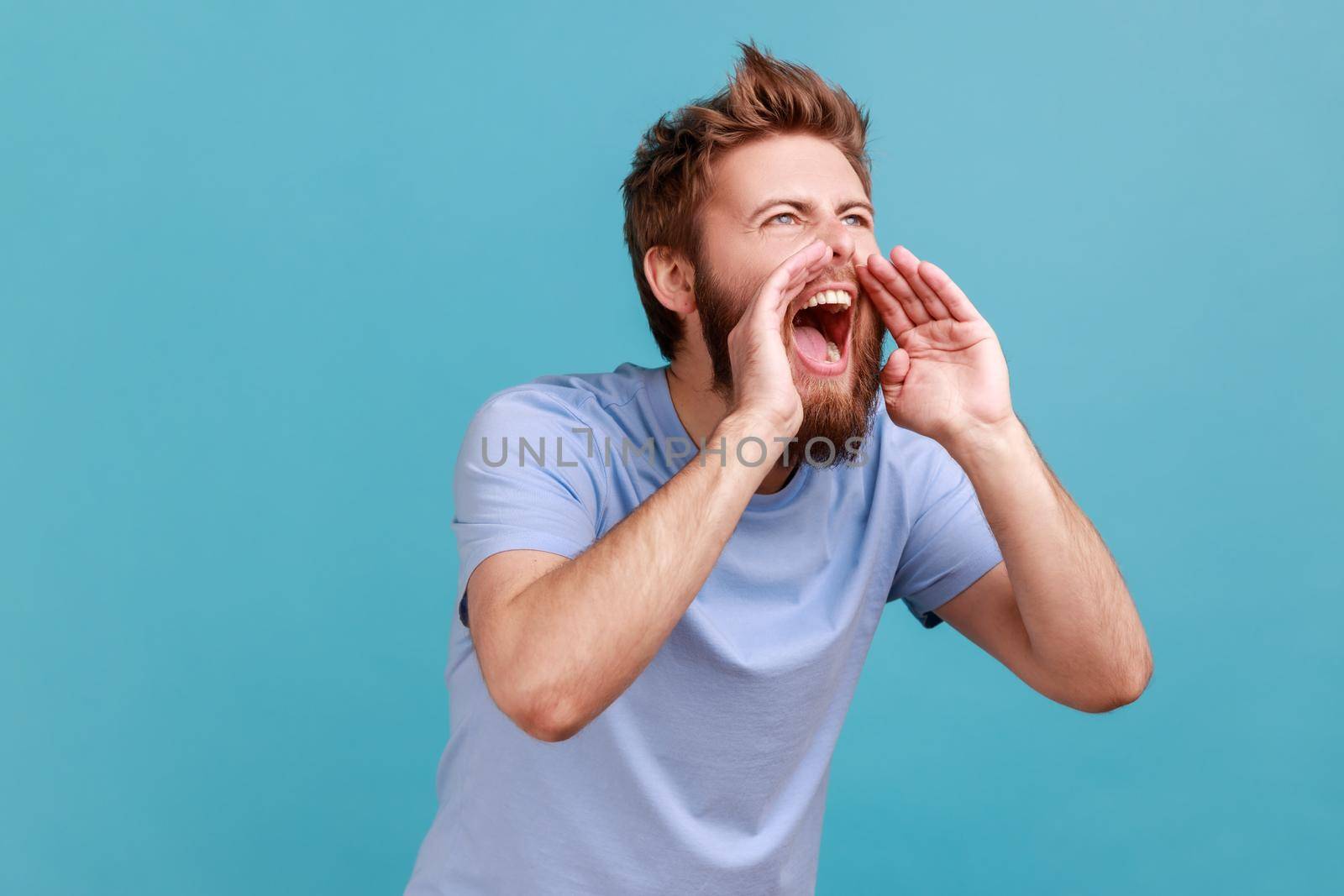 Portrait of angry handsome bearded man loudly screaming widely opening mouth holding hands near mouth, yelling announcing his opinion. Indoor studio shot isolated on blue background.