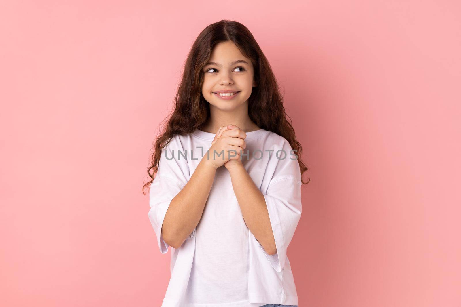 Portrait of smiling happy little girl wearing white T-shirt thinking, dreaming with joyous smiling face, pleasant fantasy, positive hopes. Indoor studio shot isolated on pink background.