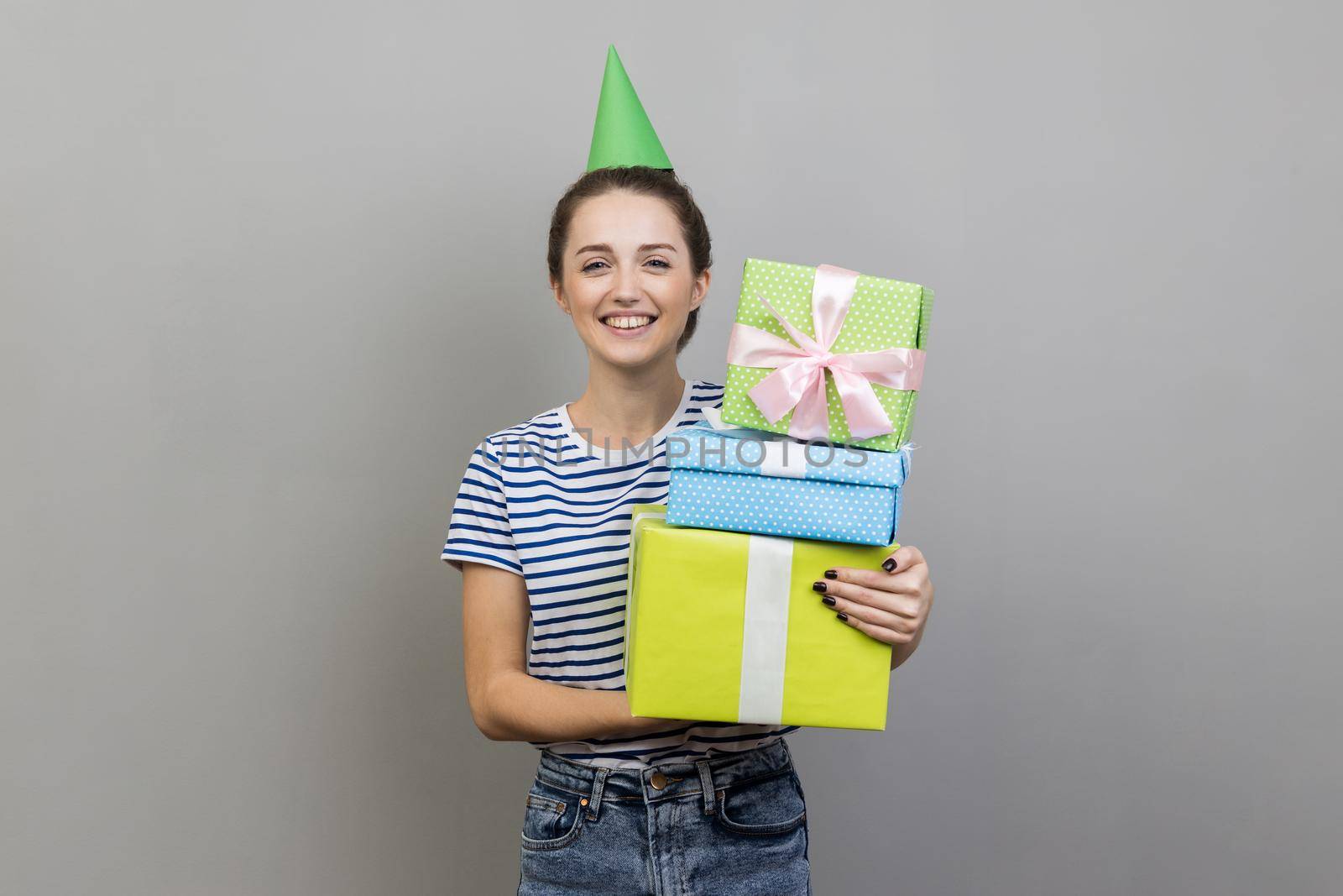 Portrait of smiling woman wearing striped T-shirt and party cone, holding stack of present boxes, celebrating birthday. Indoor studio shot isolated on gray background.
