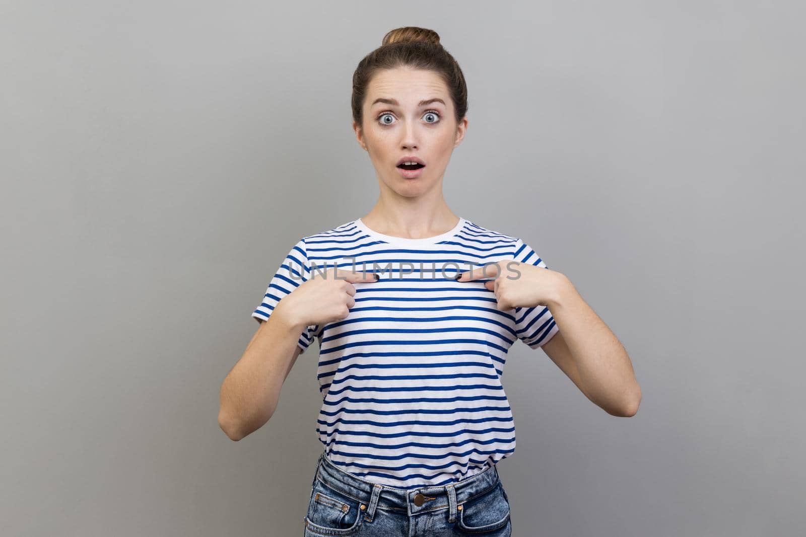 Portrait of astonished woman wearing striped T-shirt pointing at herself, asks who me, has surprised expression, shocked being picked. Indoor studio shot isolated on gray background.