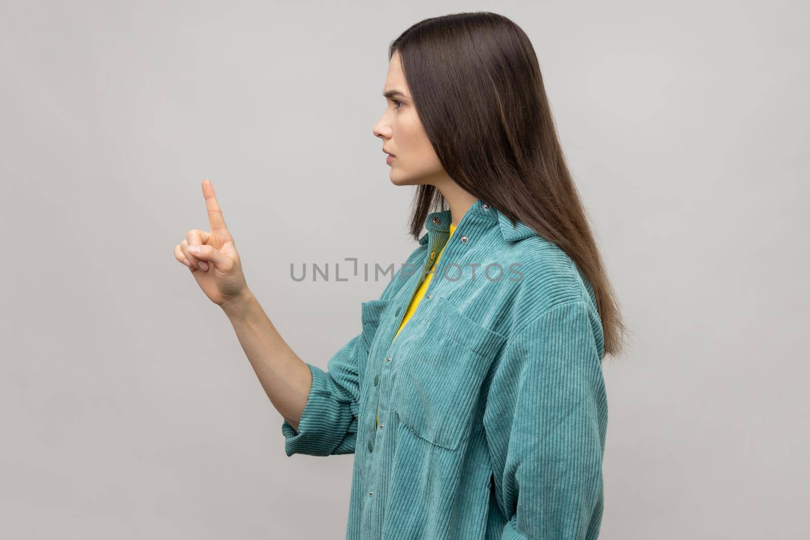 Be careful. Side view of strict bossy woman looking at camera seriously, pointing finger and warning, wearing casual style jacket. Indoor studio shot isolated on gray background.