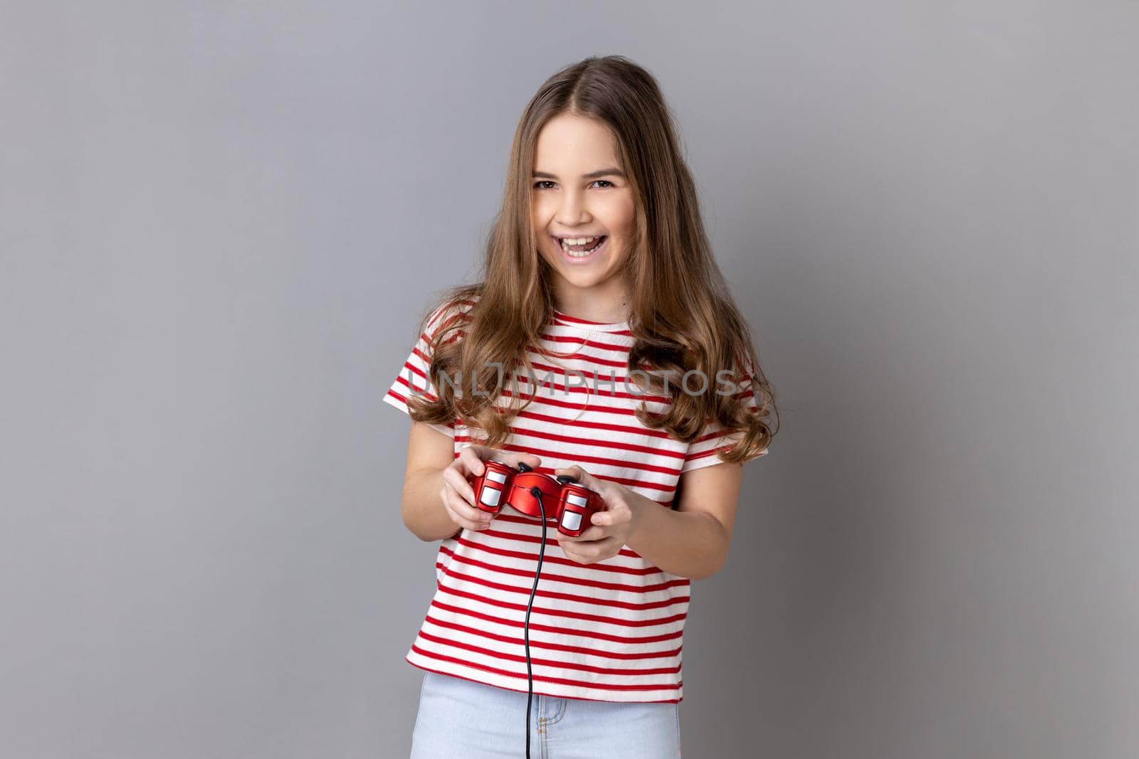 Portrait of little girl wearing striped T-shirt holding in hands red gamepad joystick, looking at camera with optimistic expression, enjoying game. Indoor studio shot isolated on gray background.