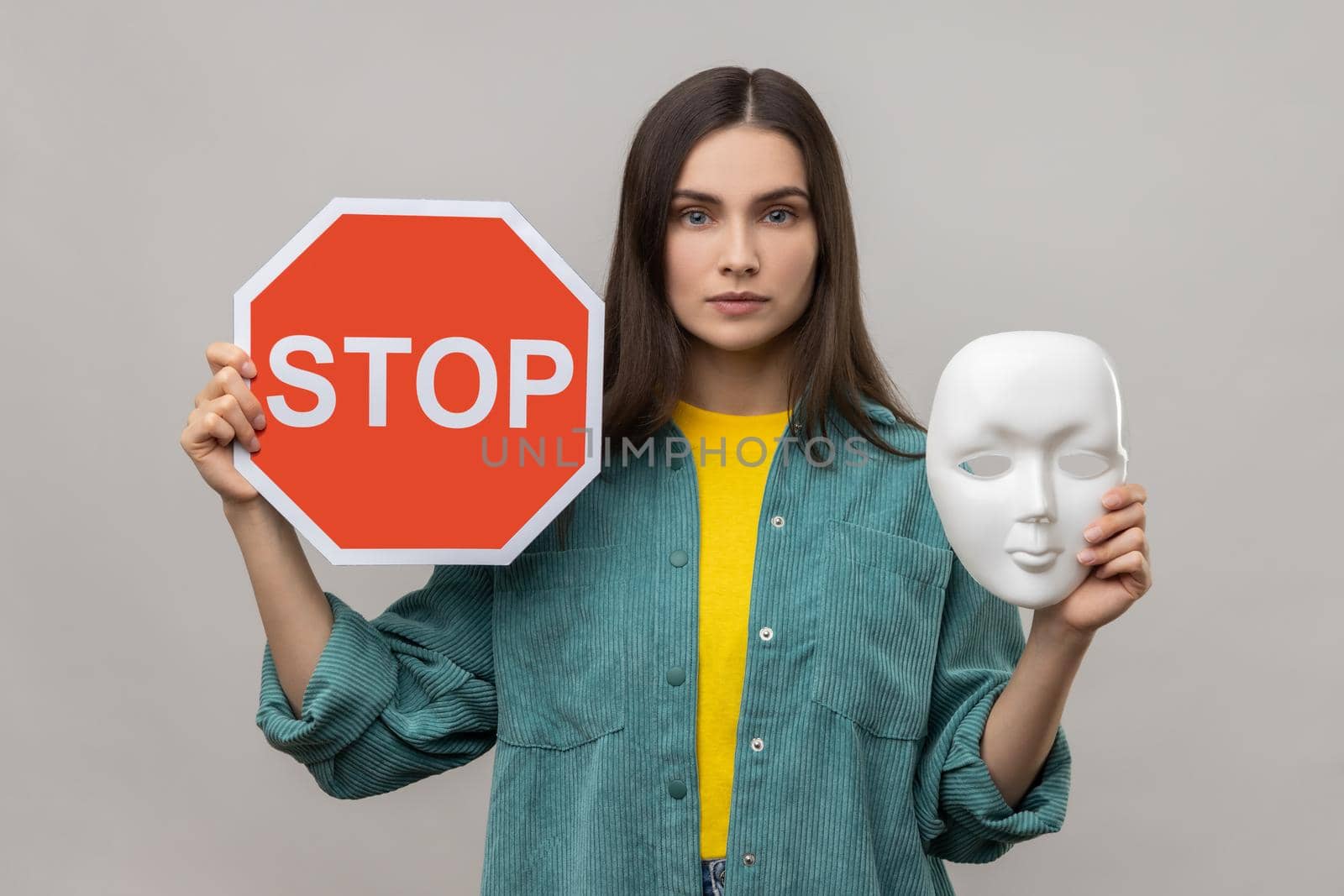 Portrait of serious dark haired woman holding red stop sign and white mask, looking at camera with strict expression, wearing casual style jacket. Indoor studio shot isolated on gray background.