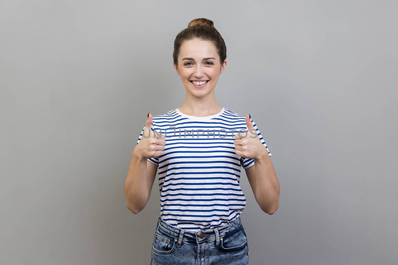 Portrait of cute joyful woman wearing striped T-shirt standing and looking at camera with toothy smile, showing thumb up, like gesture. Indoor studio shot isolated on gray background.