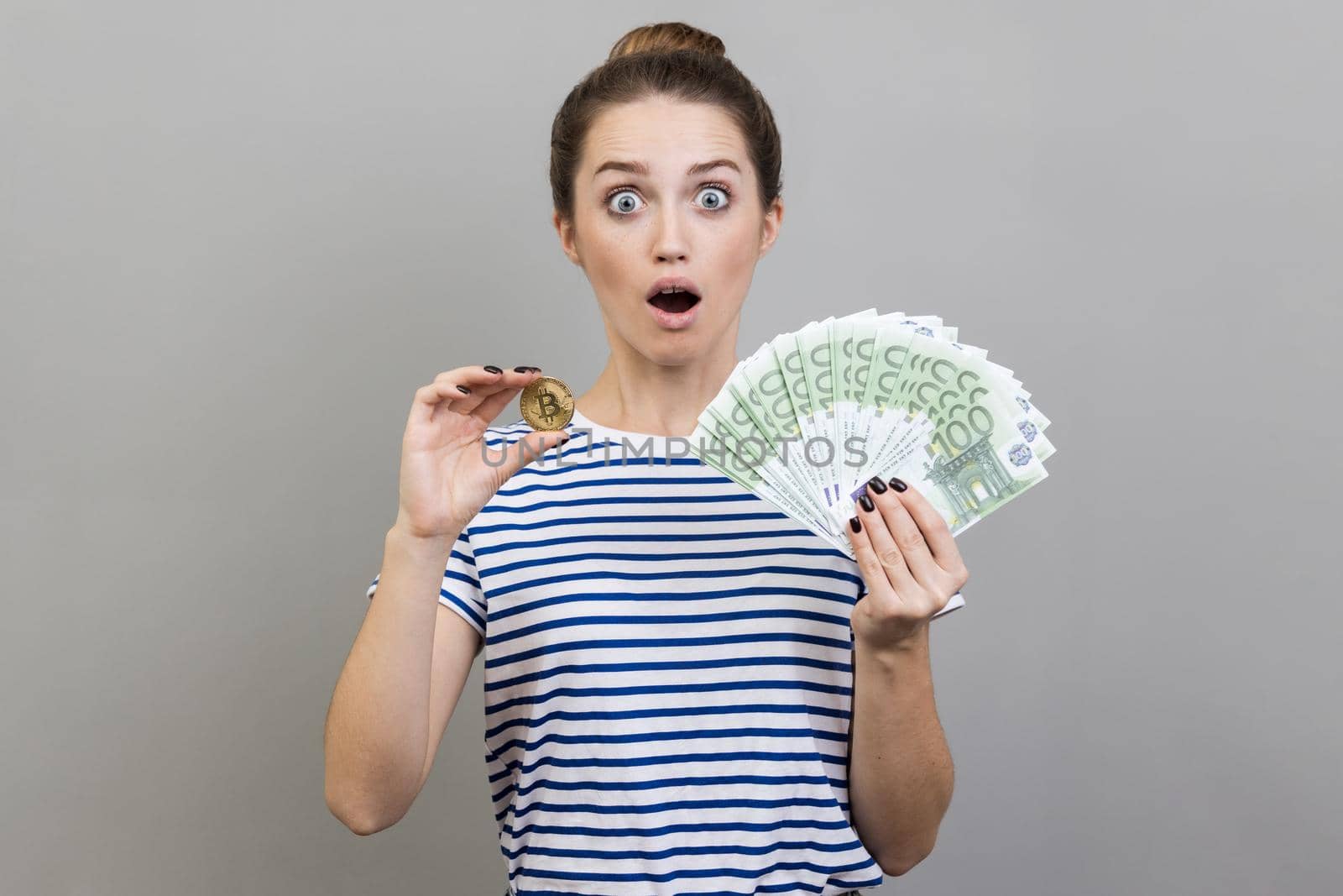 Portrait of surprised amazed shocked woman wearing striped T-shirt holding golden coin of crypto currency and big fan of euro banknotes. Indoor studio shot isolated on gray background.