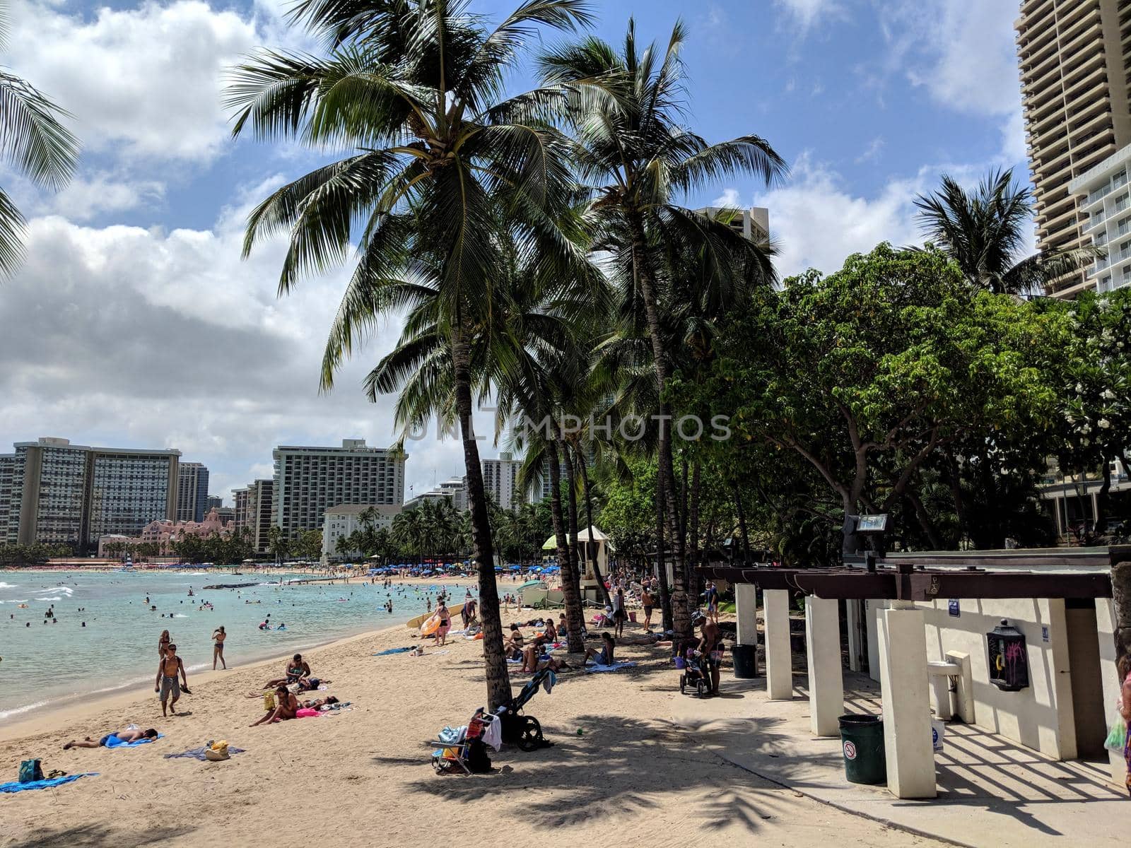 Waikiki, Hawaii - July 19, 2018: People play in the protected water and hang out by bathrooms on the beach in world famous tourist area Waikiki on a beautiful day with hotels in the distance. 