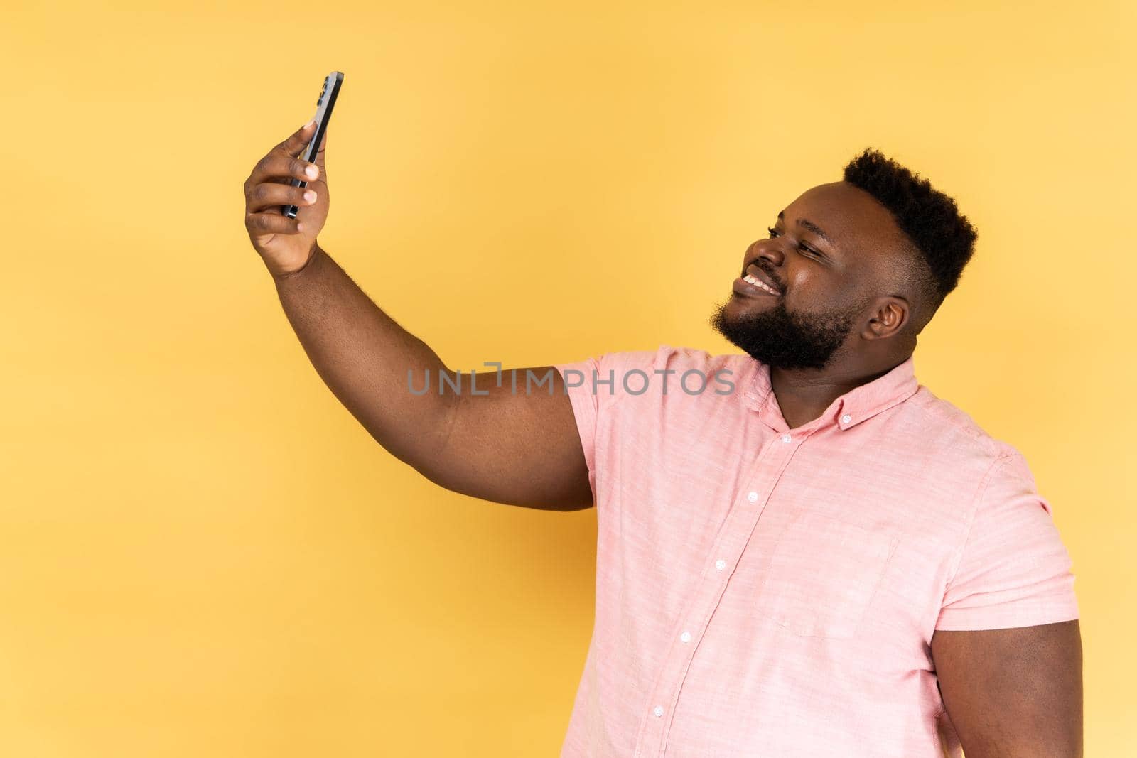 Portrait of positive optimistic man wearing pink shirt talking on video call, having online conversation on mobile phone, taking selfie. Indoor studio shot isolated on yellow background.