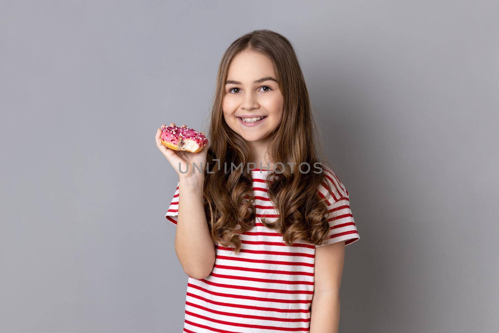 Portrait of delighted happy adorable little girl wearing striped T-shirt delicious sugary donut and smiling genuinely to camera. Indoor studio shot isolated on gray background.