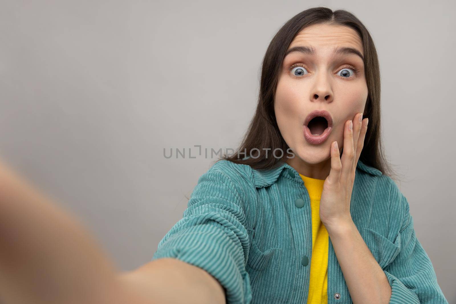 Surprised woman taking selfie with shocked facial expression, looking with big eyes and open mouth, POV, wearing casual style jacket. Indoor studio shot isolated on gray background.